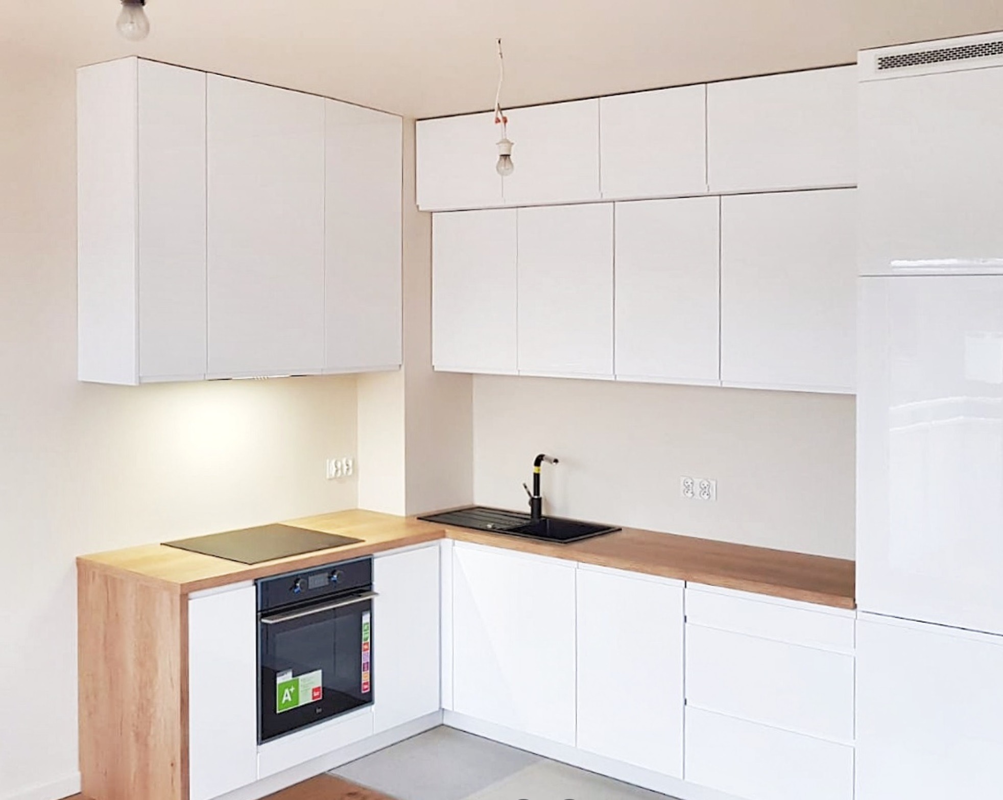 Nowoczesna kuchnia z białymi, lakierowanymi frontami szafek, drewnianym blatem i czarnym zlewem oraz płytą indukcyjną, widok na wiszące szafki i oświetlenie podszafkowe.