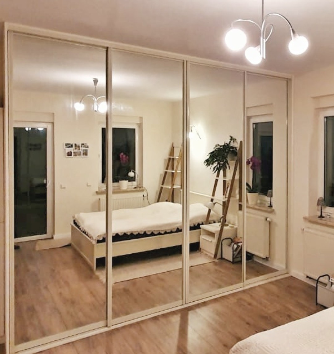 Duża szafa z lustrzanymi przesuwnymi drzwiami, odbijająca wnętrze sypialni z łóżkiem, drabiną dekoracyjną i roślinami w doniczkach na parapecie.