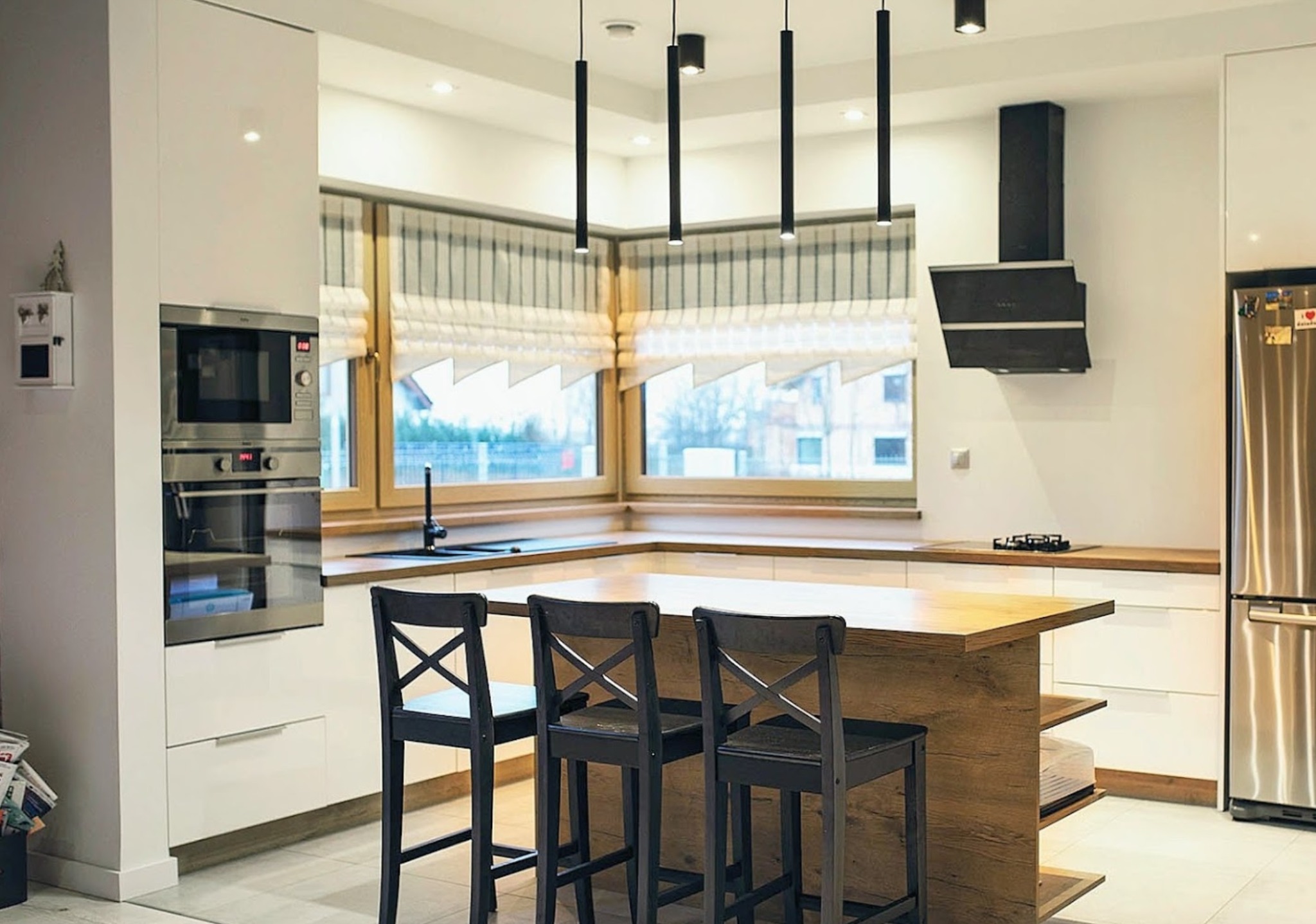 Nowoczesna kuchnia z wyspą, białe szafki z połyskiem, drewniany blat wyspy i okienny, czarne hokery z krzyżowym oparciem, minimalistyczne lampy wiszące, okno z roletami rzymskimi.