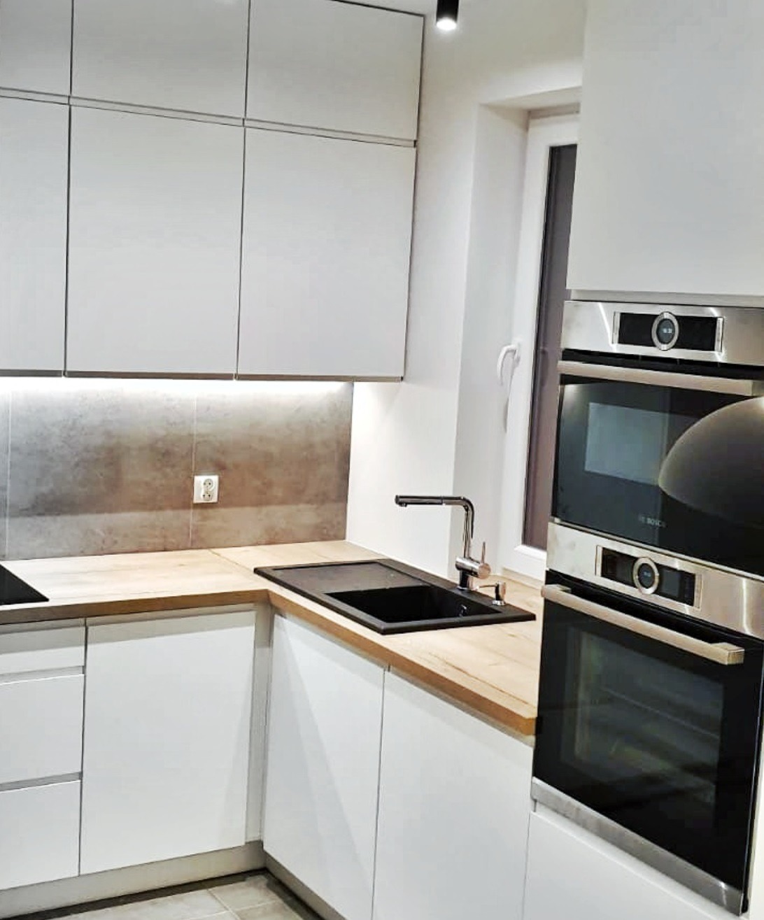 Nowoczesna kuchnia z białymi, gładkimi frontami szafek, drewnianym blatem, czarnym zlewozmywakiem i dwoma piekarnikami marki Bosch w zabudowie.