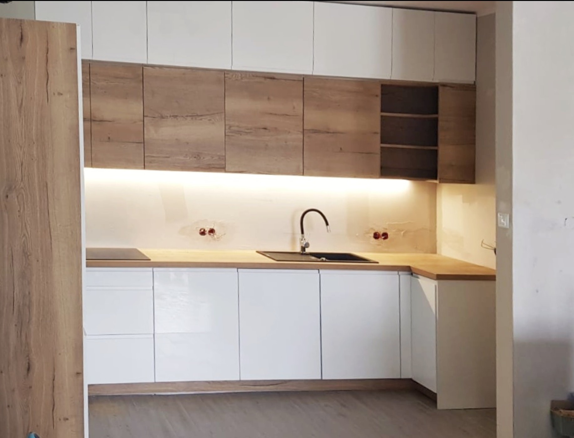 Nowoczesna kuchnia z białymi frontami dolnych szafek i drewnopodobnymi górnymi szafkami, podświetlona listwą LED, widoczne niedokończone wykończenie ściany.