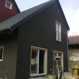 Elewacja domu jednorodzinnego pomalowana na ciemny kolor, widoczne okna z białymi ramami oraz elementy wykończeniowe w kolorze drewna, na pierwszym planie lampa grzewcza.