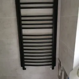 Czarny, drabinkowy grzejnik łazienkowy zamontowany na jasnych, kwadratowych płytkach ściennych. Widoczna biała głowica termostatyczna i fragment wzorzystej mozaiki w prawym dolnym rogu.