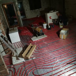 Robert Bielecki - Czerwone rury ogrzewania podłogowego rozłożone na srebrnej folii, w tle pracownik przy białym styropianie, widoczne narzędzia, drabina i kartony, surowy stan wykończenia wnętrza.