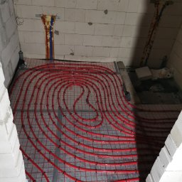 Robert Bielecki - Czerwone rury ogrzewania podłogowego ułożone na srebrnej macie w surowym wnętrzu, widoczne fragmenty ścian z białego bloczka i instalacje na ścianie.