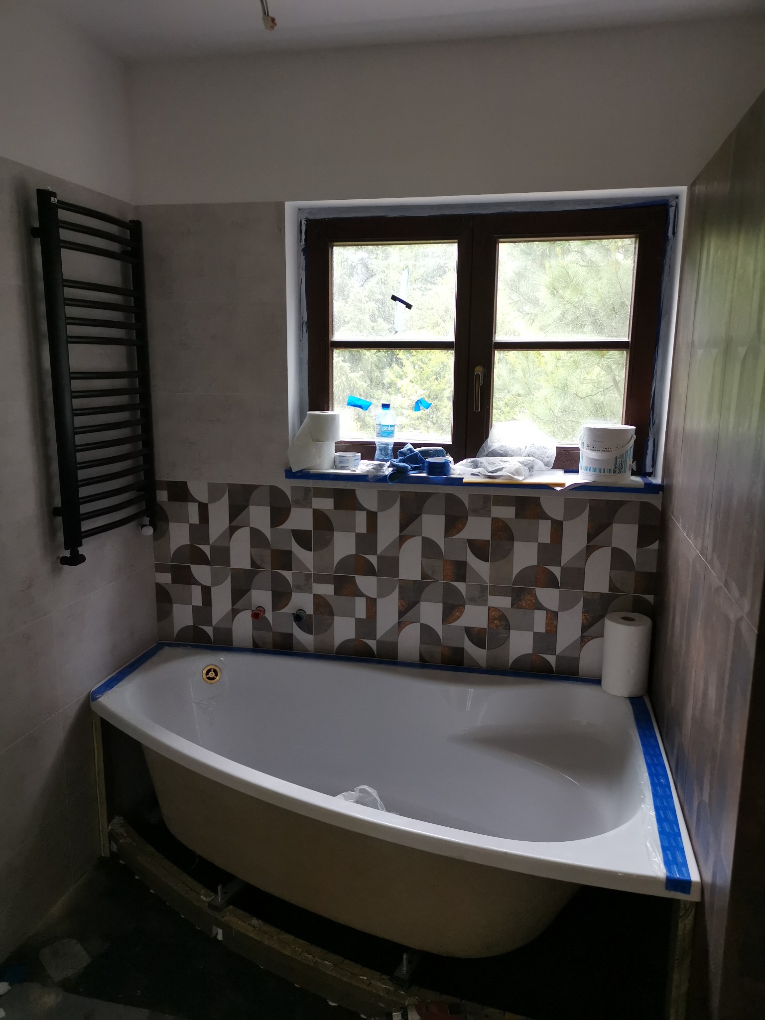 Narożna wanna z zabezpieczeniem taśmą malarską, grzejnik drabinkowy na ścianie, okno z widokiem na zieleń, płytki dekoracyjne na ścianie, butelka wody i akcesoria malarskie na parapecie, etap prac...