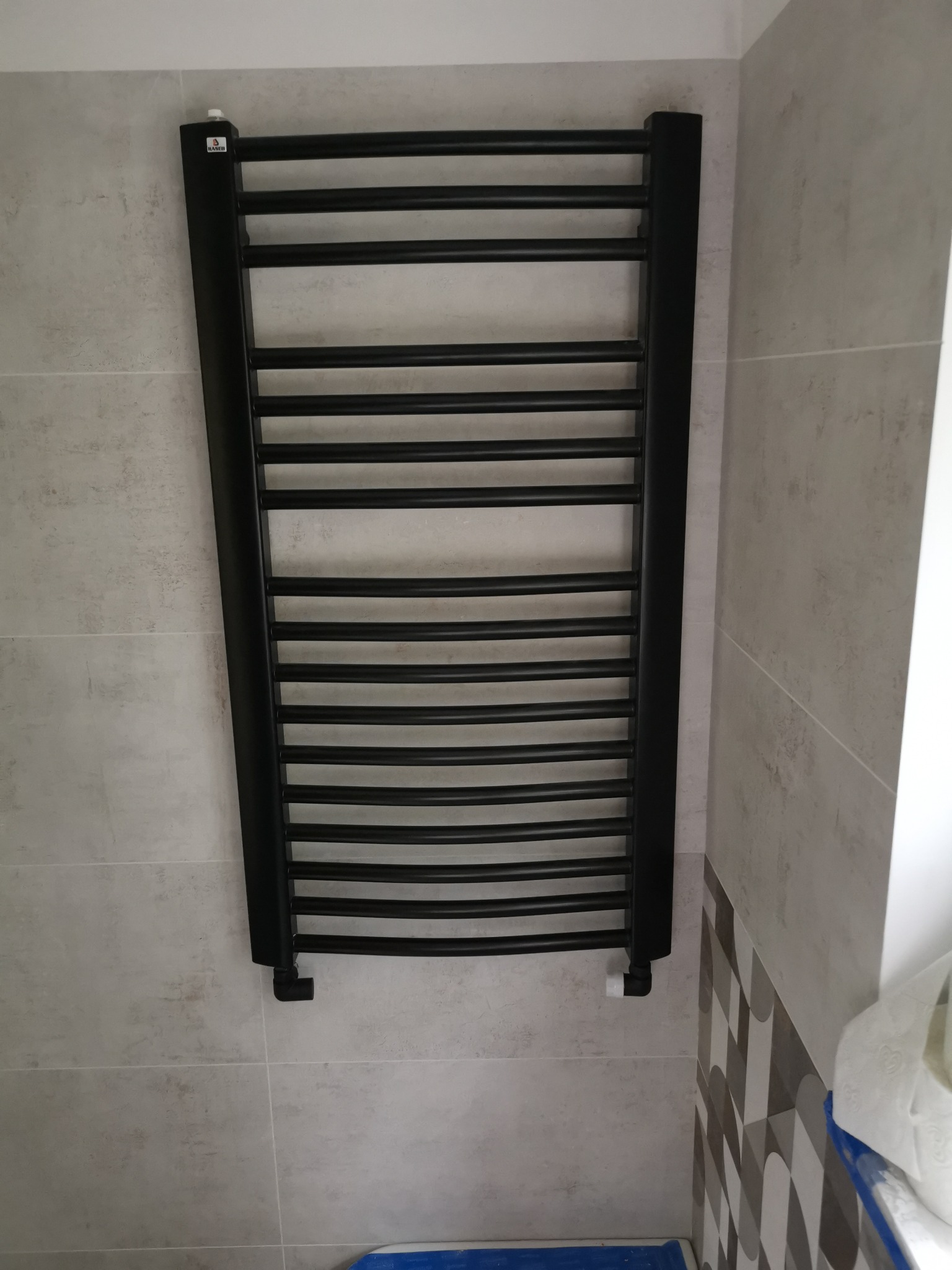 Czarny, drabinkowy grzejnik łazienkowy zamontowany na jasnych, kwadratowych płytkach ściennych. Widoczna biała głowica termostatyczna i fragment wzorzystej mozaiki w prawym dolnym rogu.