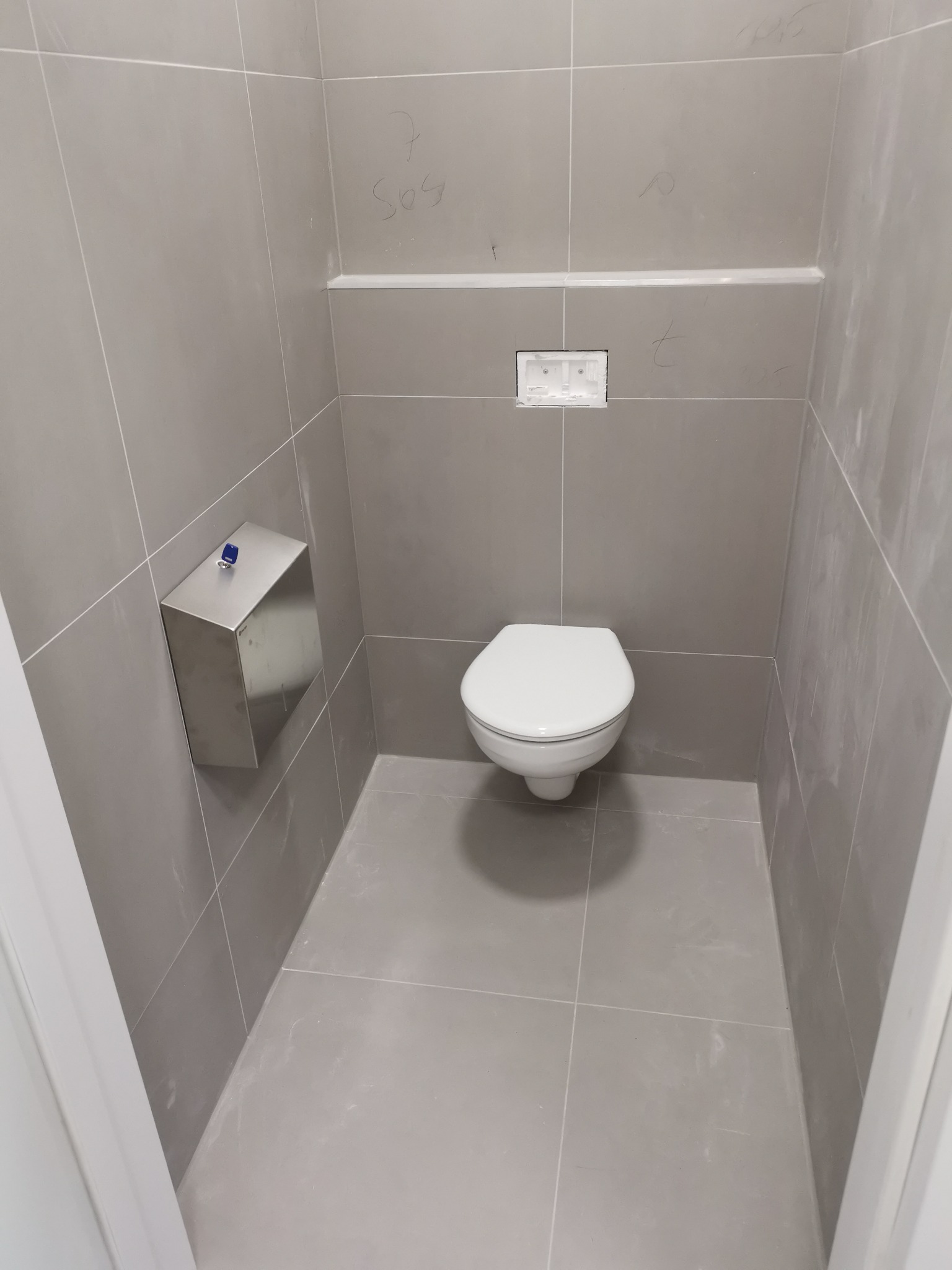 Wnętrze łazienki w trakcie remontu z zamontowaną podtynkową toaletą, widoczne szare płytki, zabudowa pod geberit i podajnik na ręczniki papierowe, ślady po ołówku na ścianach.