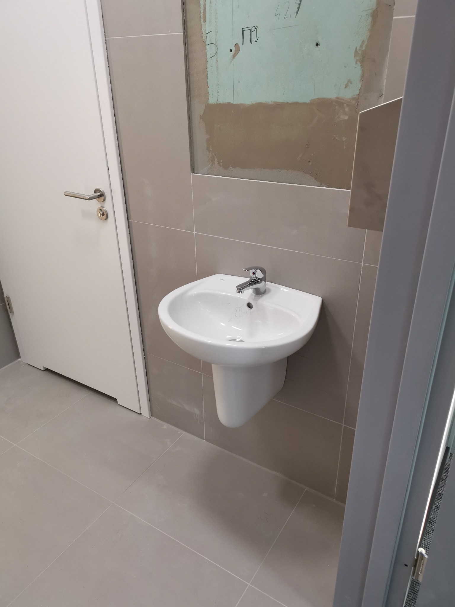 Biała umywalka ceramiczna na postumencie z chromowaną baterią zamontowana na szarej ścianie z płytkami, widoczne ślady prac wykończeniowych na ścianie, białe drzwi po lewej stronie.