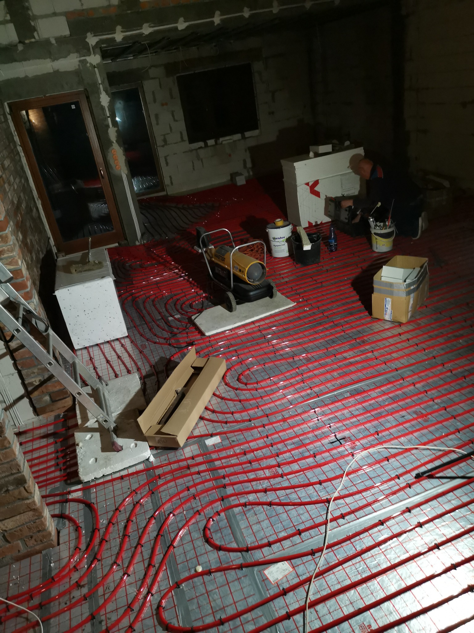 Czerwone rury ogrzewania podłogowego rozłożone na srebrnej folii, w tle pracownik przy białym styropianie, widoczne narzędzia, drabina i kartony, surowy stan wykończenia wnętrza.