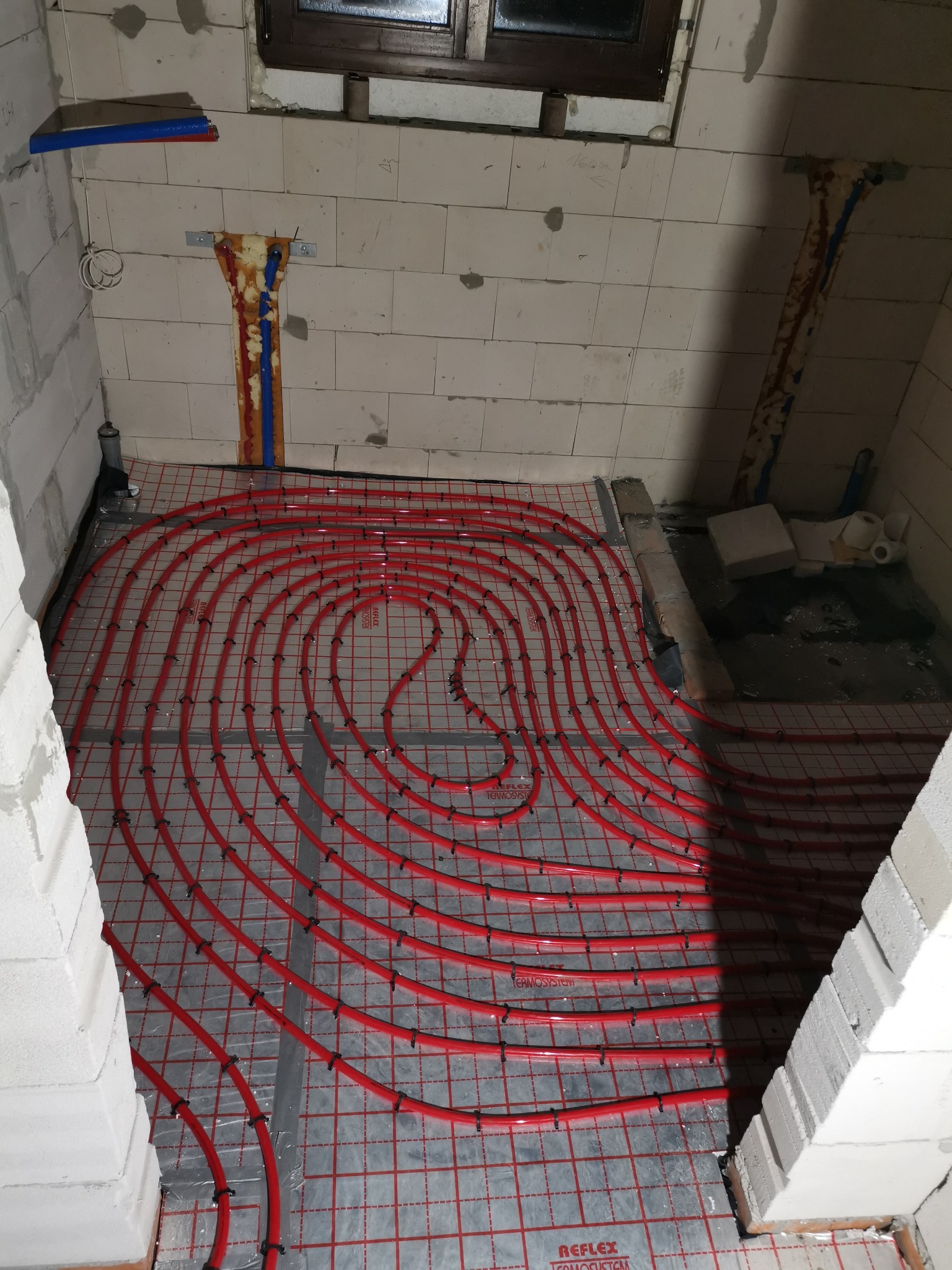 Czerwone rury ogrzewania podłogowego ułożone na srebrnej macie w surowym wnętrzu, widoczne fragmenty ścian z białego bloczka i instalacje na ścianie.