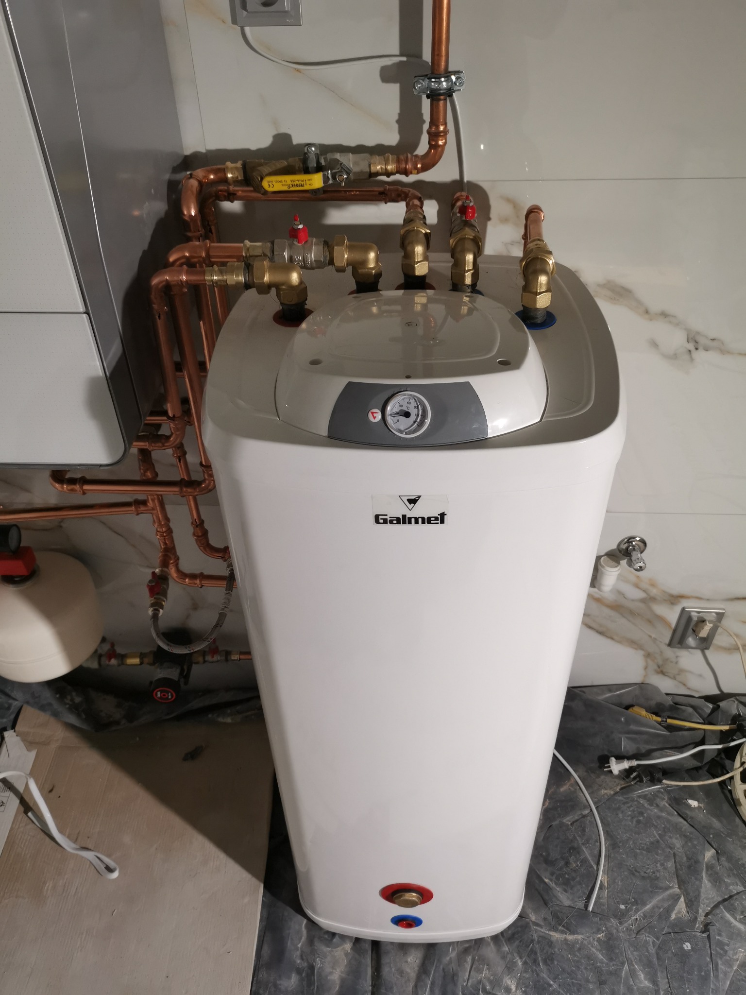Biały zasobnik ciepłej wody marki Galmet z miedzianymi rurami i zaworami, widoczny manometr, instalacja w trakcie montażu na tle marmurowej ściany.