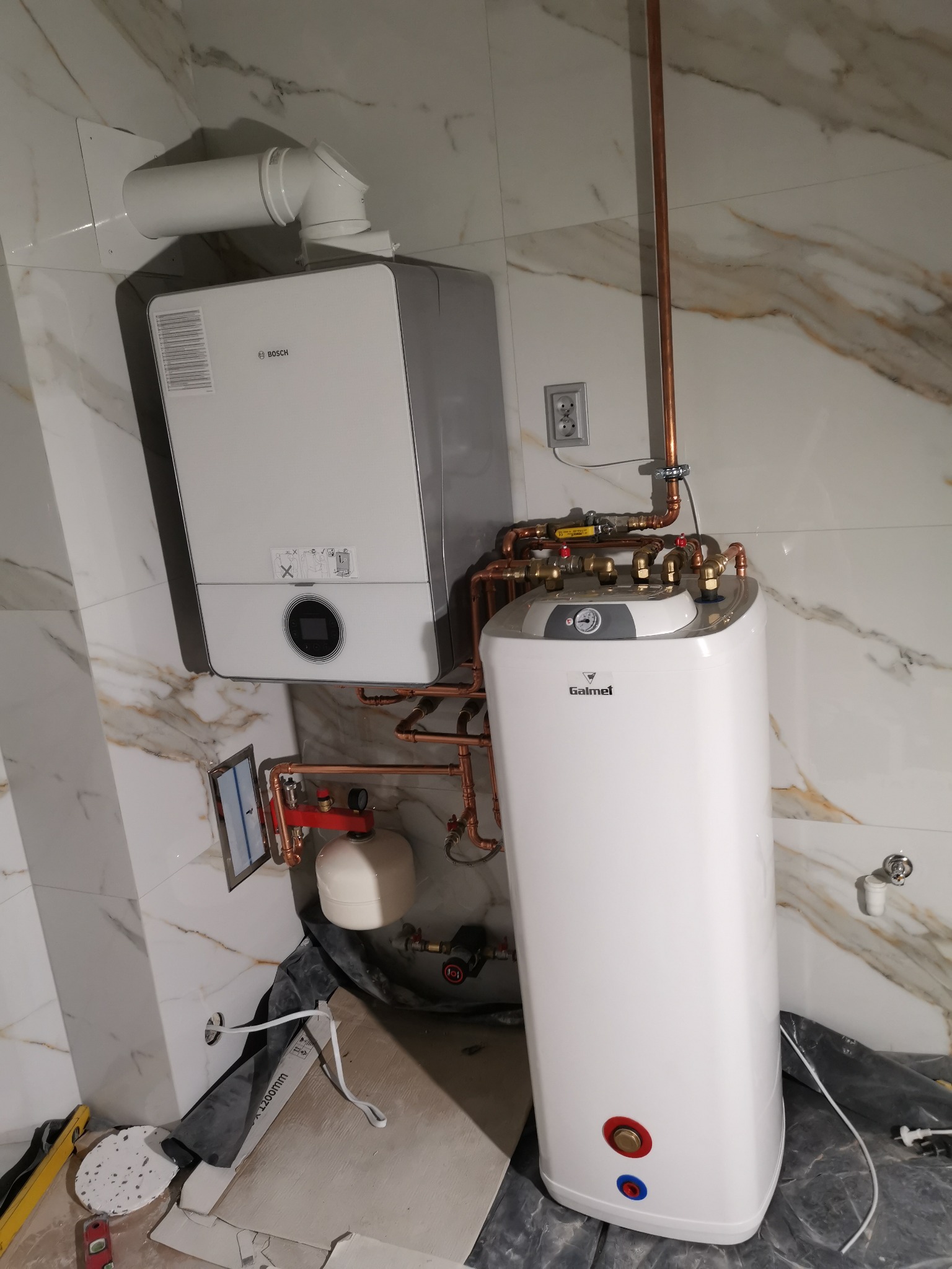 Biały, wiszący piec gazowy Bosch i stojący zasobnik Galmet z miedzianymi rurami, zamontowane na ścianie z marmurowymi płytkami, w trakcie instalacji, widoczne narzędzia i materiały budowlane.