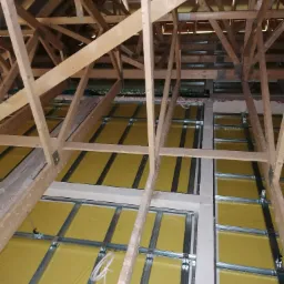 Szkielet drewniany poddasza z widoczną izolacją w kolorze żółtym i metalowymi profilami przygotowanymi do montażu płyt gipsowo-kartonowych, widok z dołu.