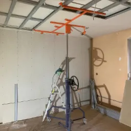 Montaż płyt na metalowym stelażu sufitowym za pomocą podnośnika; widoczna drabina z wkrętarką i częściowo wykończona ściana.