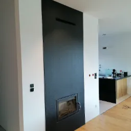 Nowoczesny kominek w obudowie z czarnej płyty, wkomponowany w białą ścianę, z widoczną częścią kuchni w tle i drewnianą podłogą.
