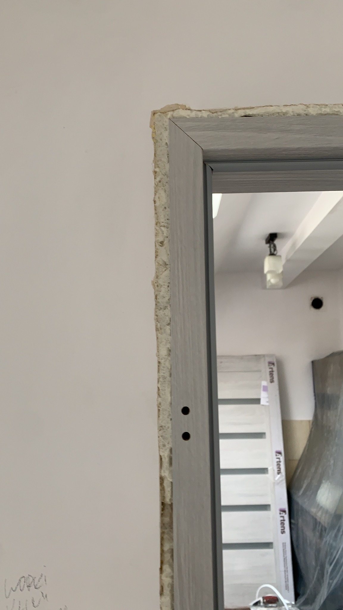 Fragment ościeżnicy drzwiowej w trakcie montażu, widoczne niedoskonałości ściany i pianka montażowa, w tle widoczna nowa, zapakowana ościeżnica.