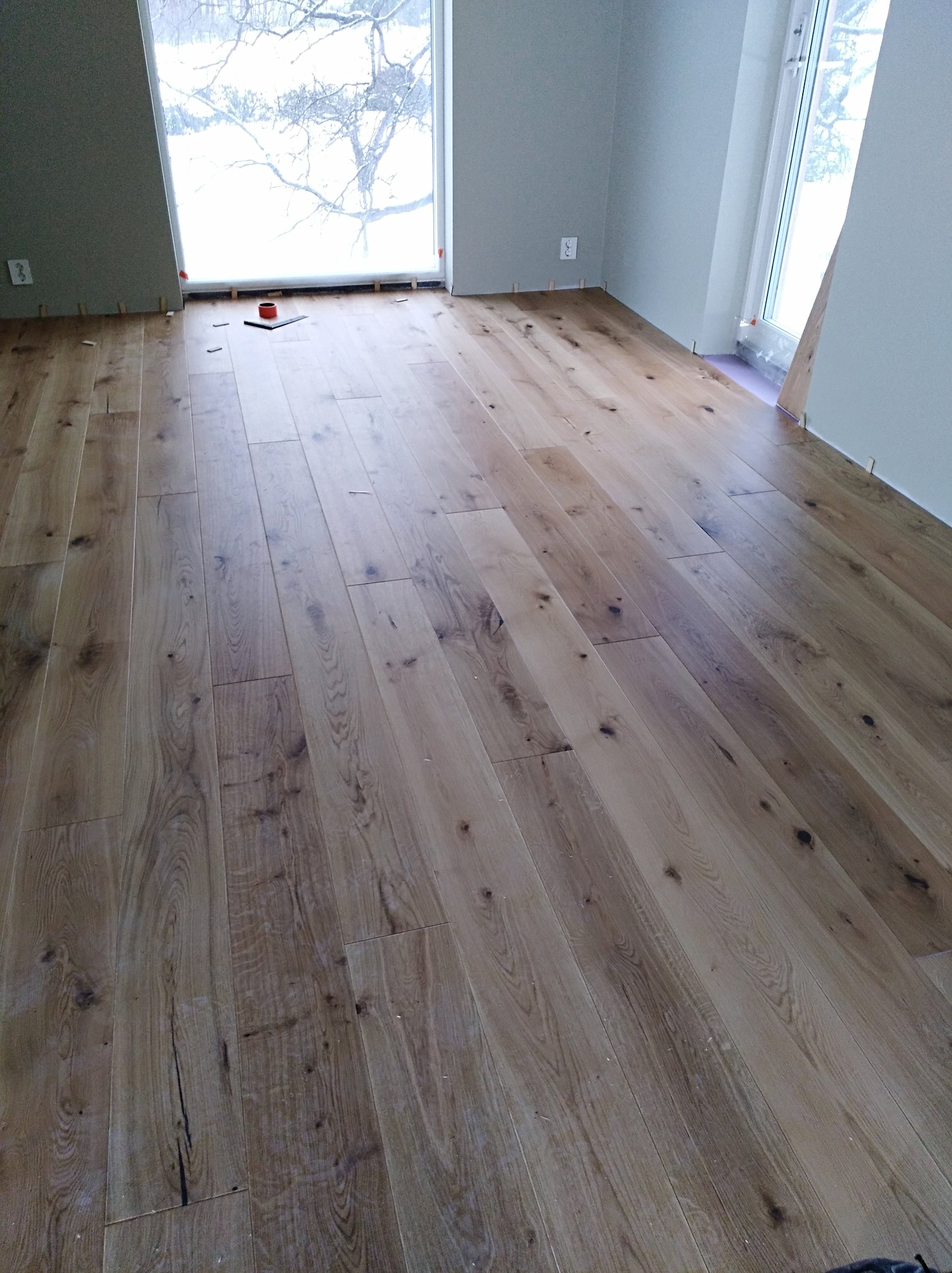 Nowo ułożony parkiet z jasnego drewna w pustym pokoju z widokiem na zimowy krajobraz za oknem, widoczne kliny dystansowe wzdłuż ścian.