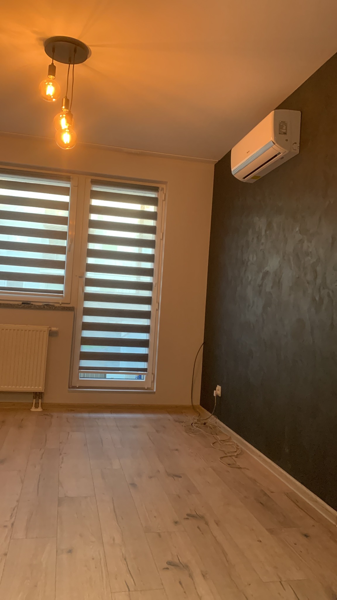 Nowoczesne wnętrze z klimatyzatorem na ciemnej ścianie, drewnianą podłogą, żaluzjami okiennymi i lampą z trzema żarówkami.