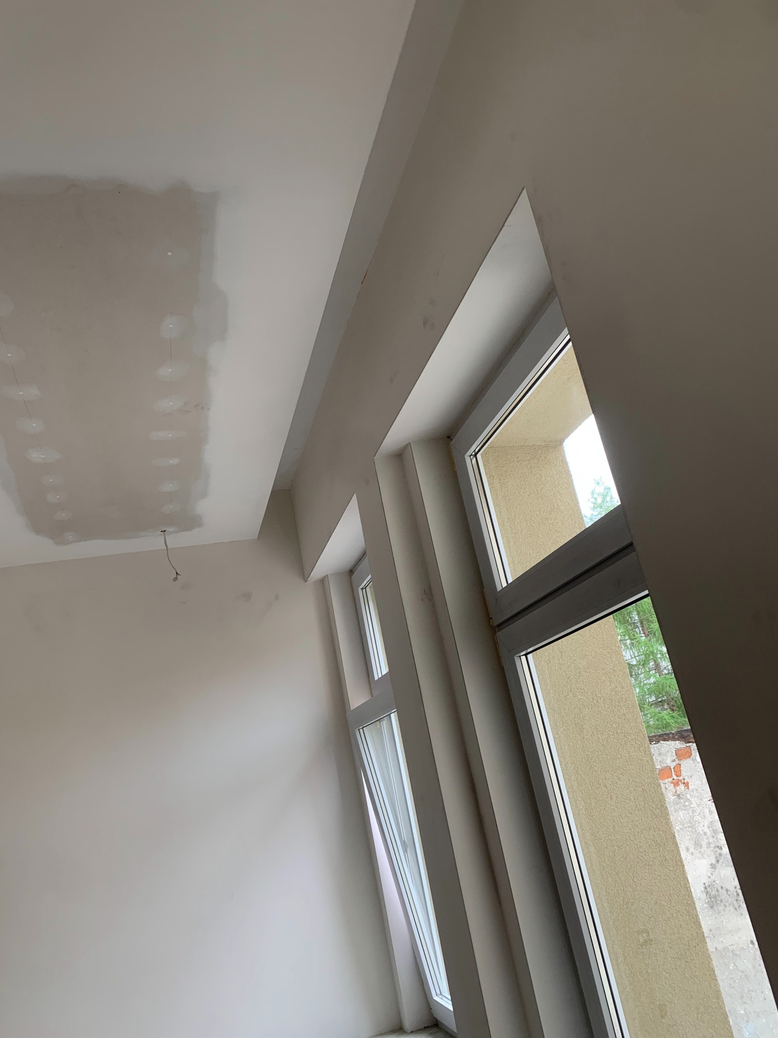 Wnętrze w trakcie remontu: sufit z widocznymi łączeniami płyt gipsowo-kartonowych przed szpachlowaniem, kabel zwisający z sufitu, ściany pomalowane na jasny kolor, okna z widokiem na zewnątrz.