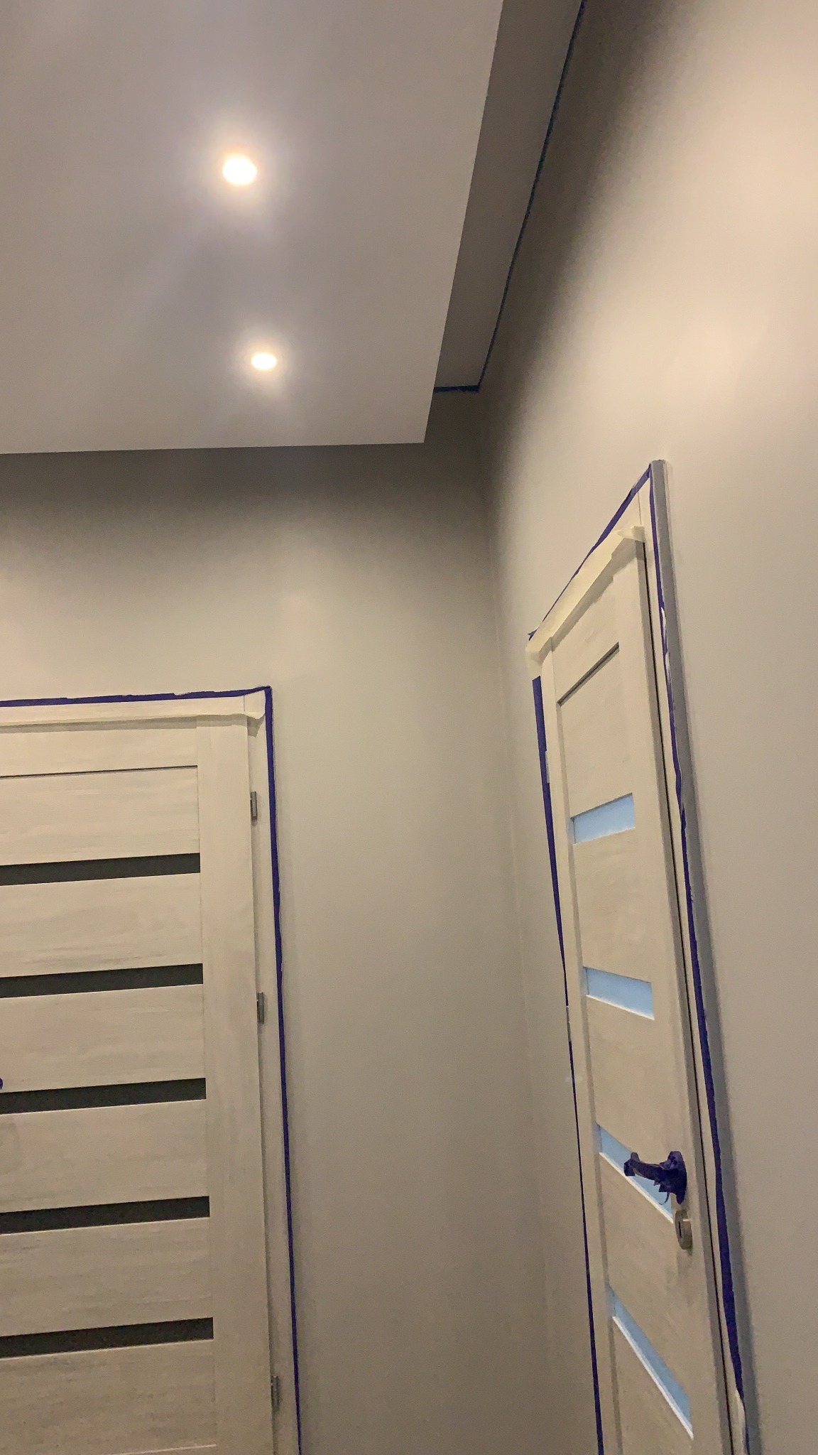 Przygotowanie do malowania ścian i sufitu: dwie białe, nowoczesne drzwi z poziomymi wstawkami zabezpieczone taśmą malarską, sufit podwieszany z dwoma punktowymi źródłami światła.