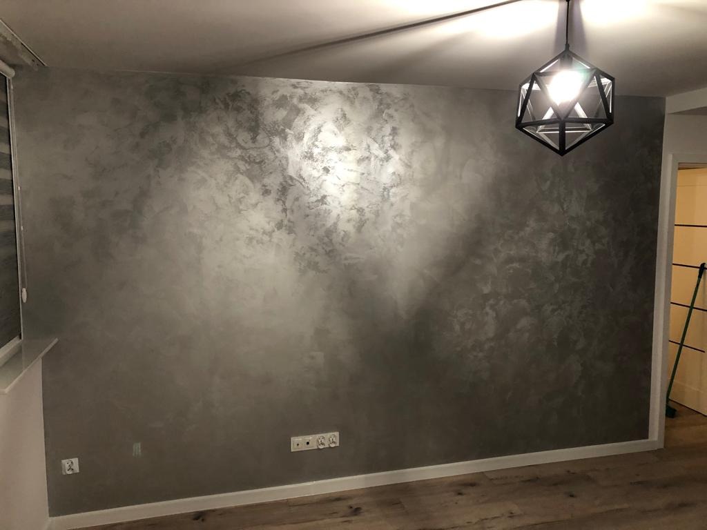 Ściana w pokoju pokryta dekoracyjną szpachlą w odcieniach szarości z efektem metalicznym, oświetlona nowoczesną lampą wiszącą z geometrycznym kloszem.