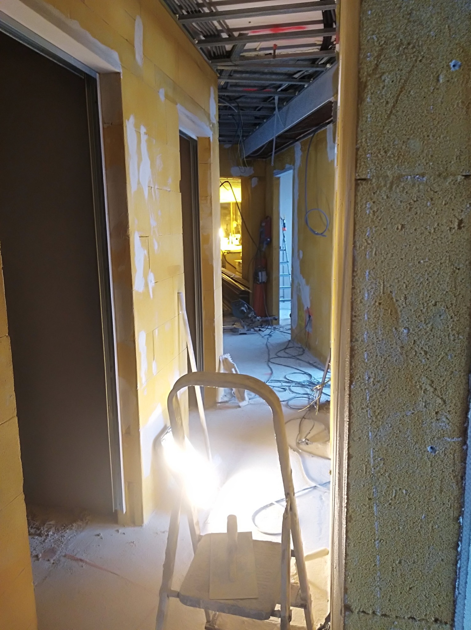 Korytarz w trakcie prac wykończeniowych: ściany z widocznymi fragmentami zaprawy, metalowa drabina z narzędziami na pierwszym planie, instalacje pod sufitem w tle.