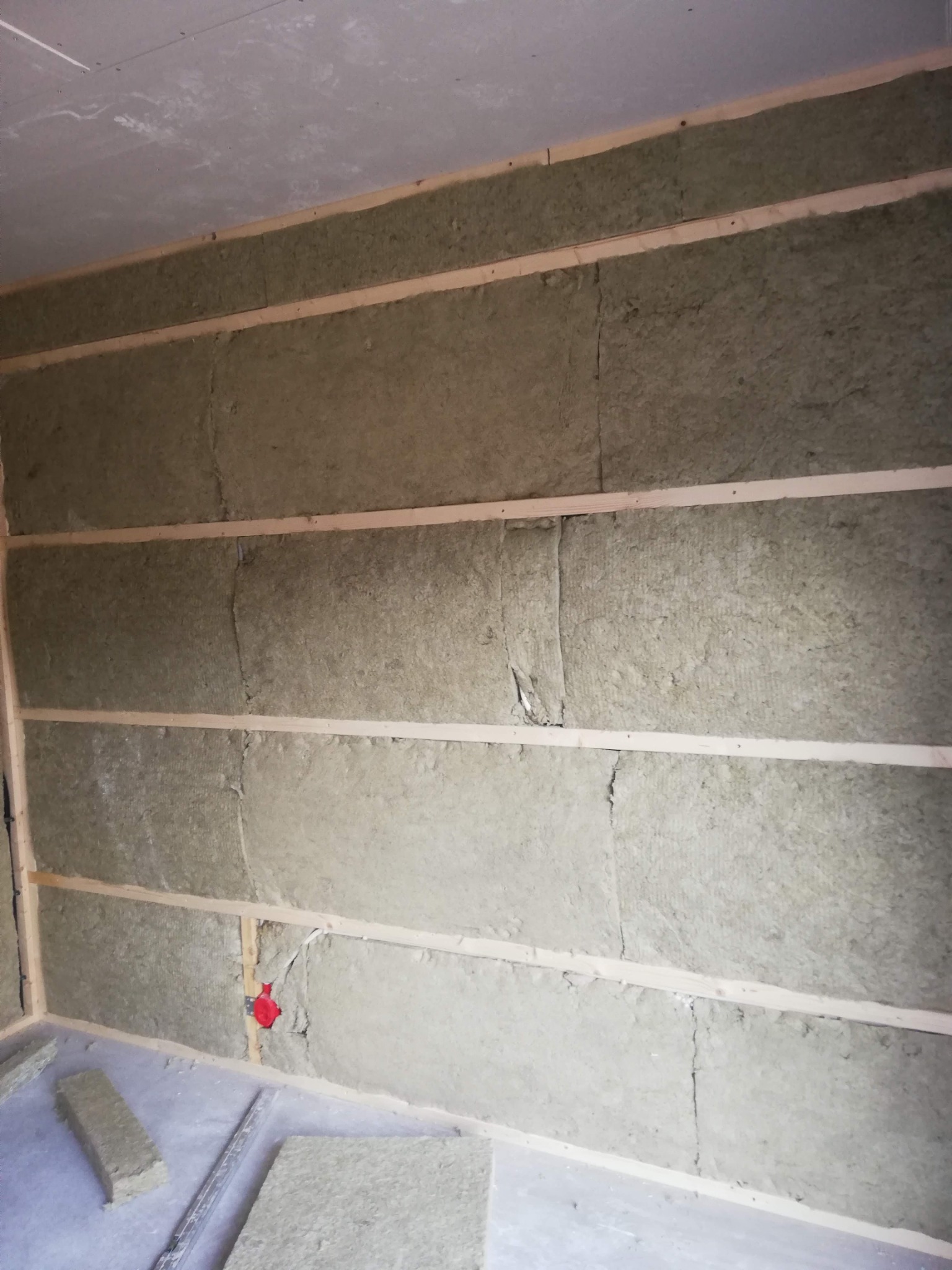 Montaż izolacji z wełny mineralnej między drewnianymi legarami na ścianie poddasza, widoczna czerwona puszka elektryczna i fragmenty izolacji na podłodze.