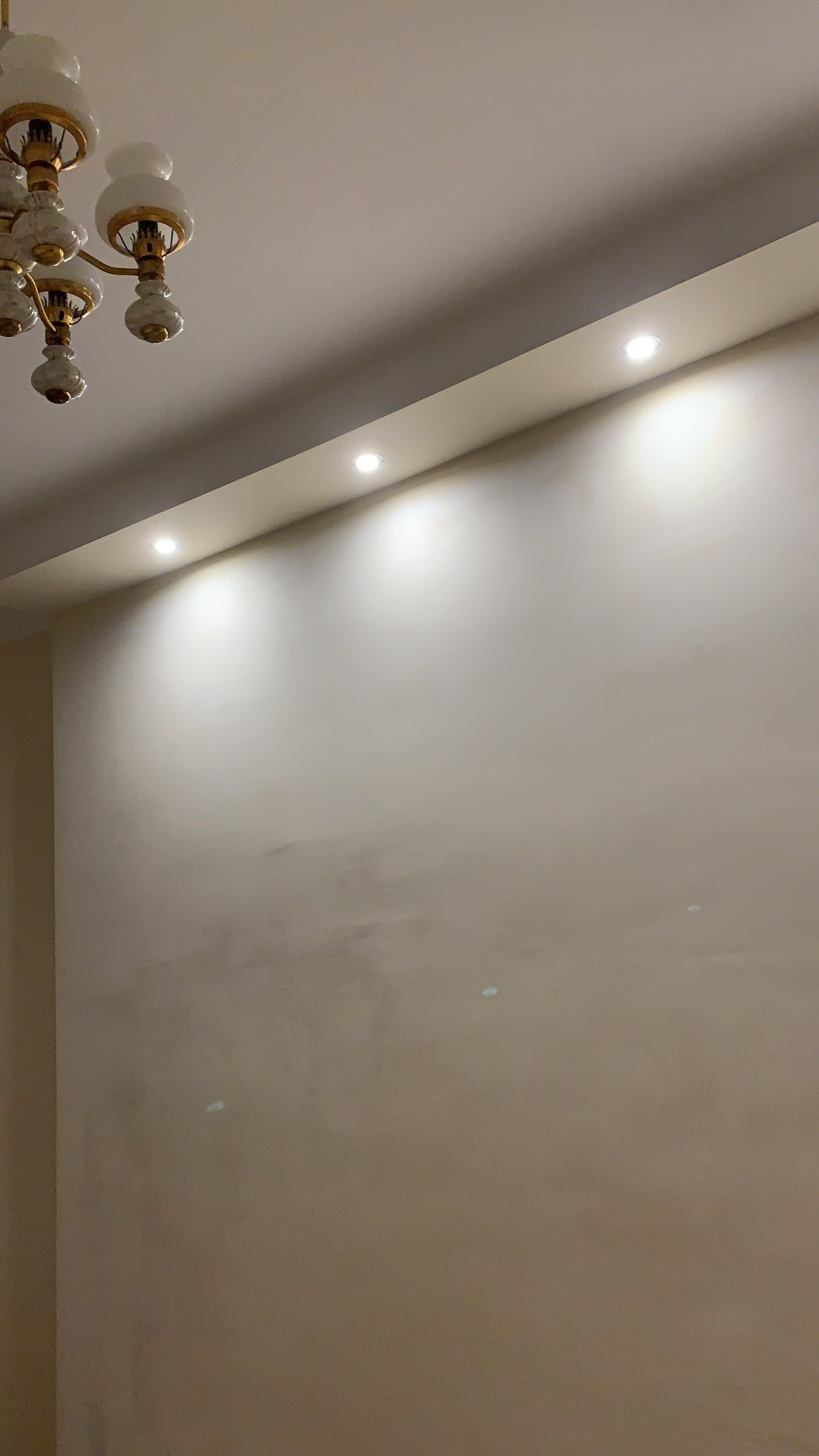 Ściana w jasnym odcieniu beżu z widocznymi śladami szpachlowania, oświetlona punktowymi lampami w suficie i klasycznym żyrandolem z białymi kloszami.