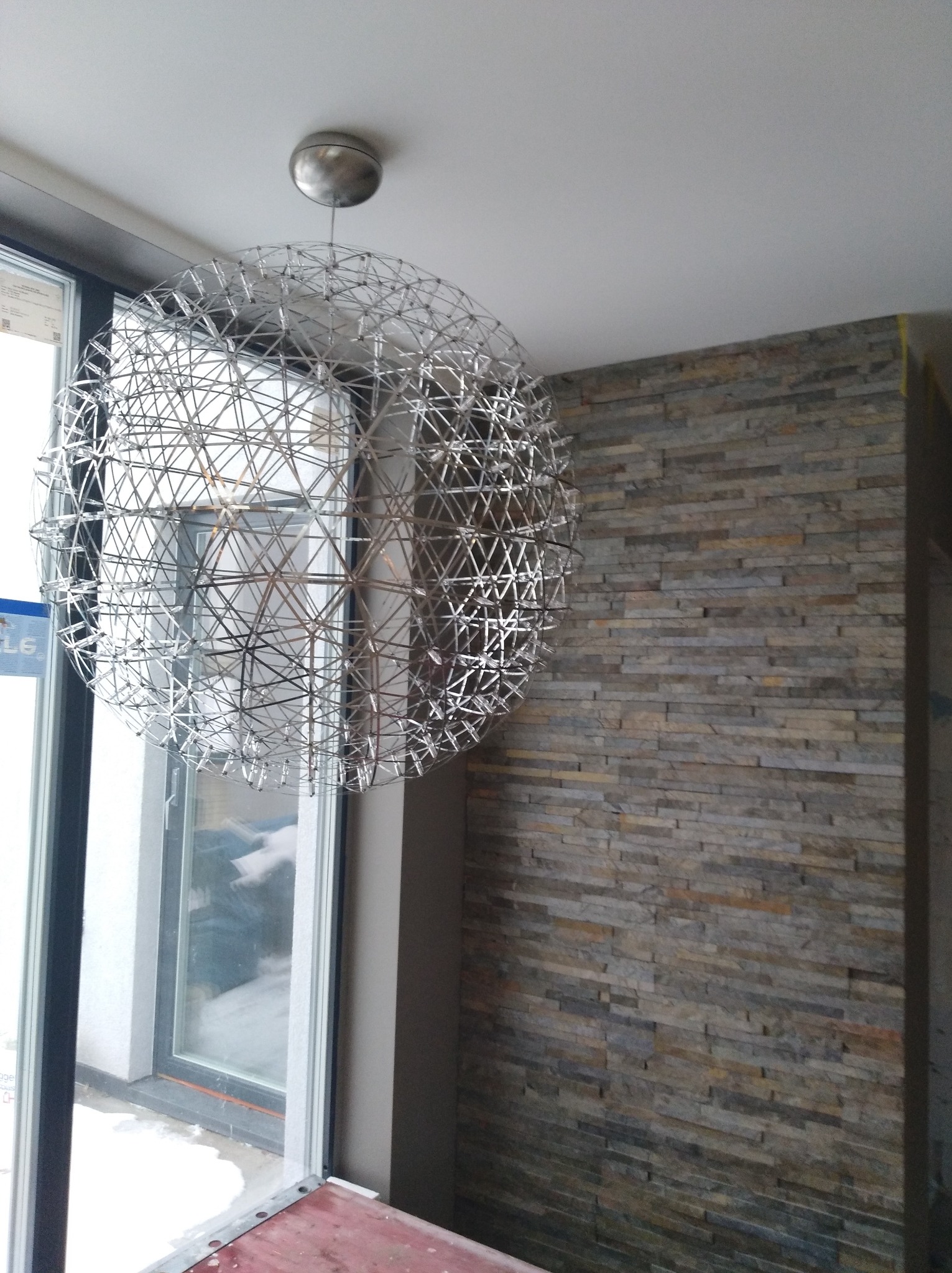 Nowoczesna lampa wisząca o ażurowej, kulistej konstrukcji z metalowych prętów, na tle ściany wyłożonej wąskimi, kamiennymi płytkami w odcieniach szarości i brązu, widok z dołu.