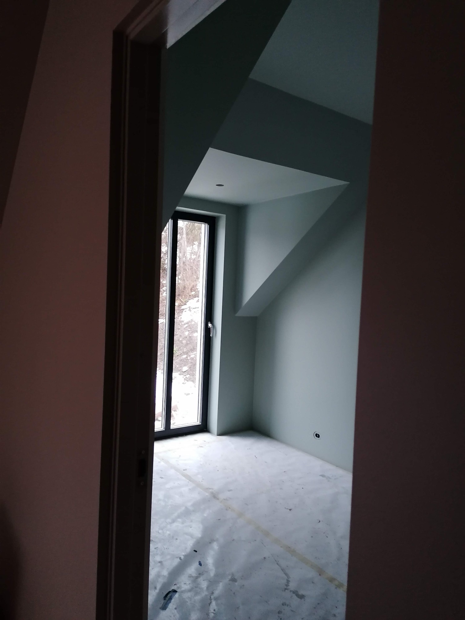 Poddasze po malowaniu na pastelowy odcień zieleni, widok przez futrynę drzwi, okno w czarnej ramie z widokiem na zaśnieżone drzewa, podłoga zabezpieczona folią.