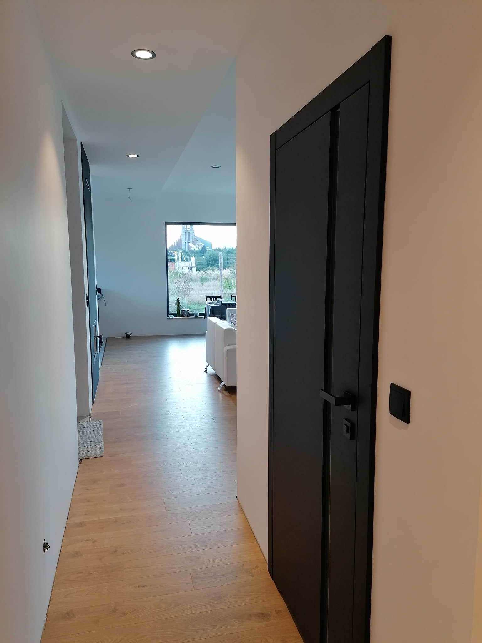 Nowoczesny korytarz z drewnianą podłogą, czarnymi drzwiami wewnętrznymi i widokiem na zewnątrz przez okno w głębi