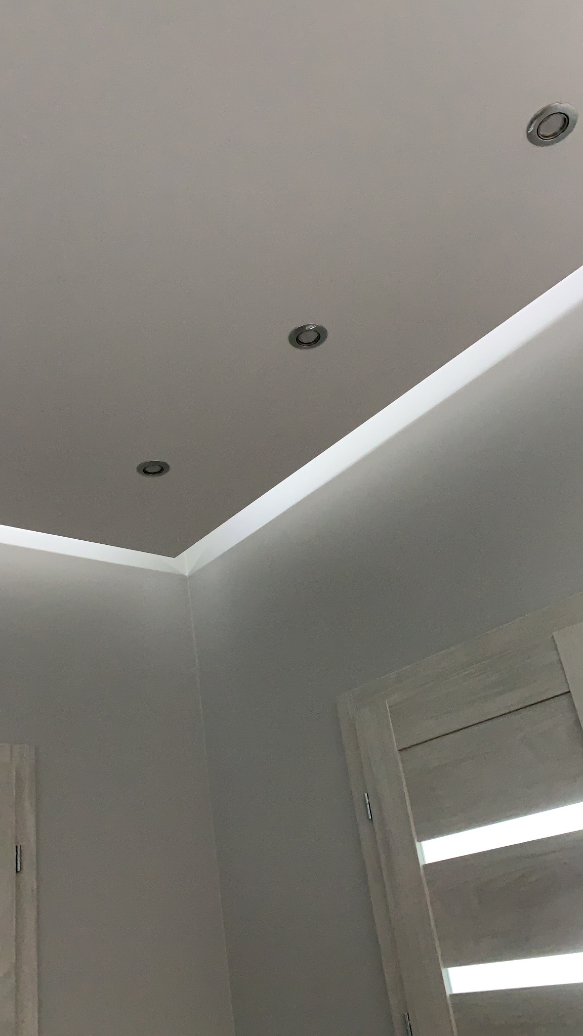 Biały sufit z trzema wbudowanymi, okrągłymi lampami halogenowymi i ozdobnym oświetleniem LED wzdłuż krawędzi, widoczne fragmenty białych drzwi.