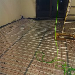 PIOTR ŁUKASZEWICZ - Rozłożone białe rury grzewcze na podłodze z czerwoną siatką, widoczny zwój rur, drabina i narzędzia instalacyjne, w tle ściana i okno.