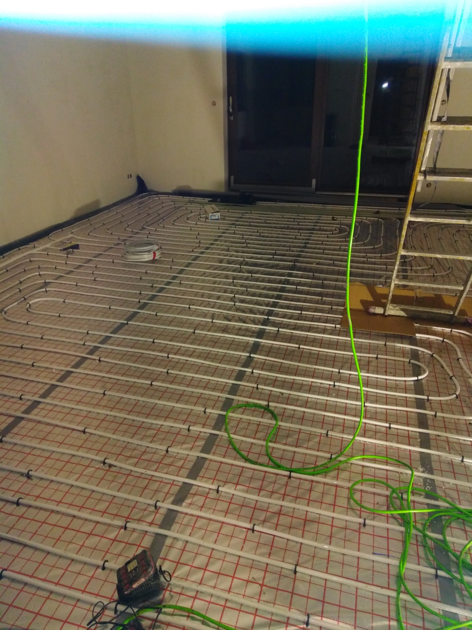 Rozłożone białe rury grzewcze na podłodze z czerwoną siatką, widoczny zwój rur, drabina i narzędzia instalacyjne, w tle ściana i okno.