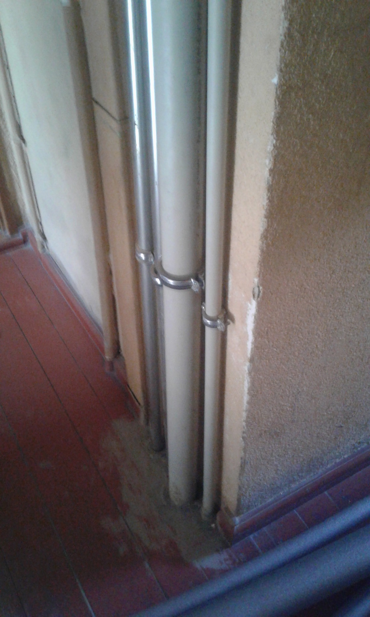 Pionowe rury, częściowo metalowe i plastikowe, zamocowane do ściany za pomocą metalowych obejm, widoczne przy podłodze z czerwonych płytek.