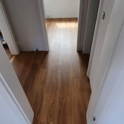 Firma ogólnobudowlana - Układanie paneli podłogowych w Gnieźnie, widok z progu na świeżo położoną, drewnopodobną podłogę w korytarzu, ściany w trakcie przygotowania do malowania, okno dachowe w tle.