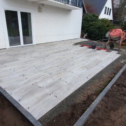 Firma ogólnobudowlana - Układanie płytek imitujących drewno na tarasie, widoczne czarne krzyżyki dystansowe, obrzeże z kostki granitowej, mieszarka do betonu w tle.