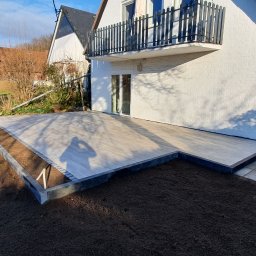 Firma ogólnobudowlana - Nowoczesny taras z jasnych desek kompozytowych, obrzeża z ciemnej kostki, świeżo nawieziona ziemia pod trawnik, cień osoby fotografującej na tarasie.