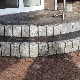 Półokrągłe, dwustopniowe schody wejściowe z kostki granitowej, prowadzące do drzwi, z kostką brukową w kolorze ceglastym wokół.