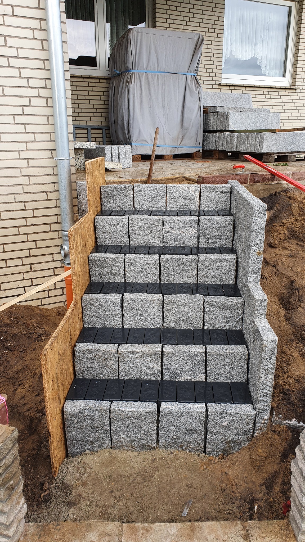 Schody zewnętrzne z kostki granitowej w trakcie budowy, widoczne czarne podkładki pod stopniami, drewniane szalunki po lewej stronie i przygotowane podłoże z ziemi po prawej.