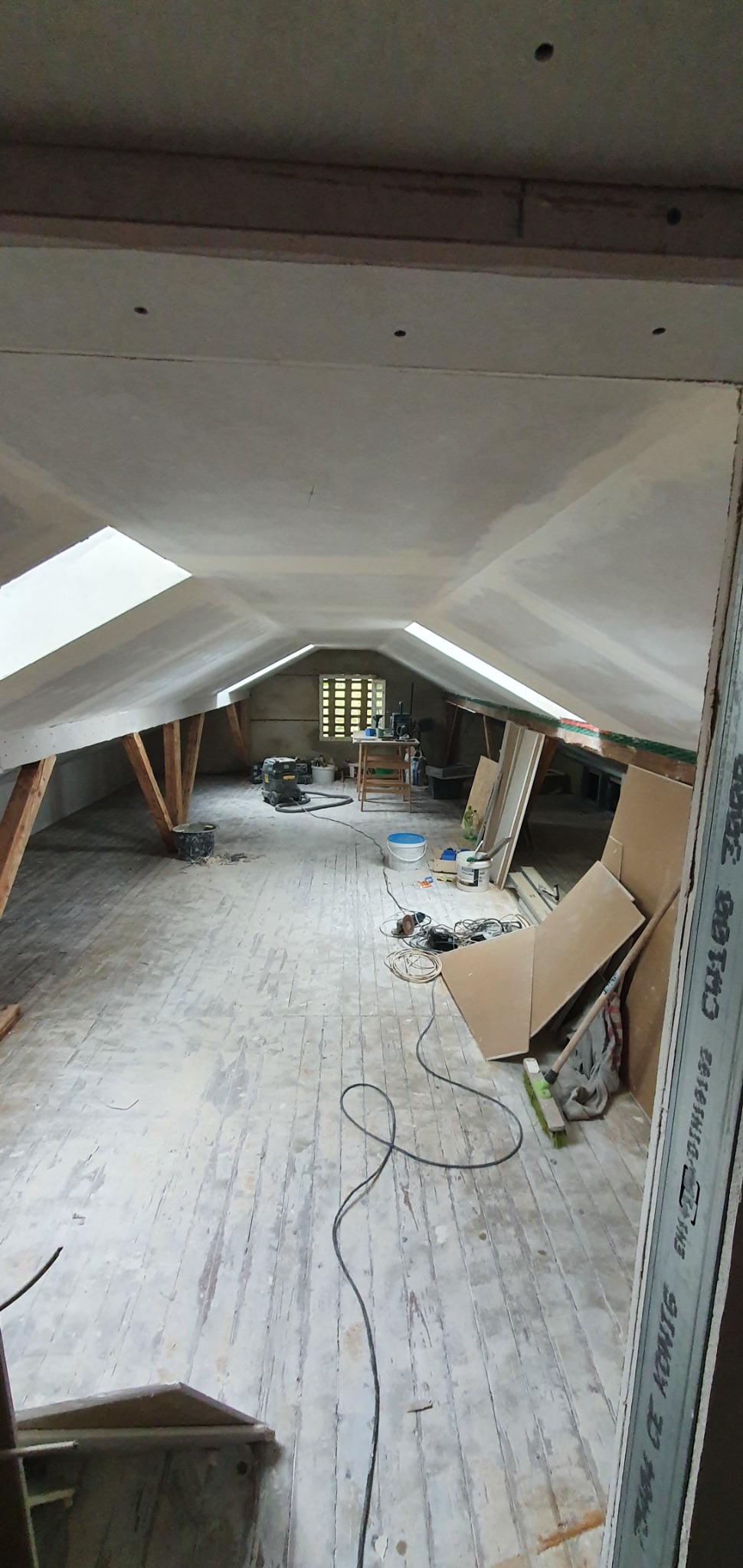 Poddasze w trakcie prac remontowych, widoczne okna dachowe, drewniane belki konstrukcyjne, płyty gipsowo-kartonowe na ścianach i suficie, oraz narzędzia i materiały budowlane na podłodze z desek.