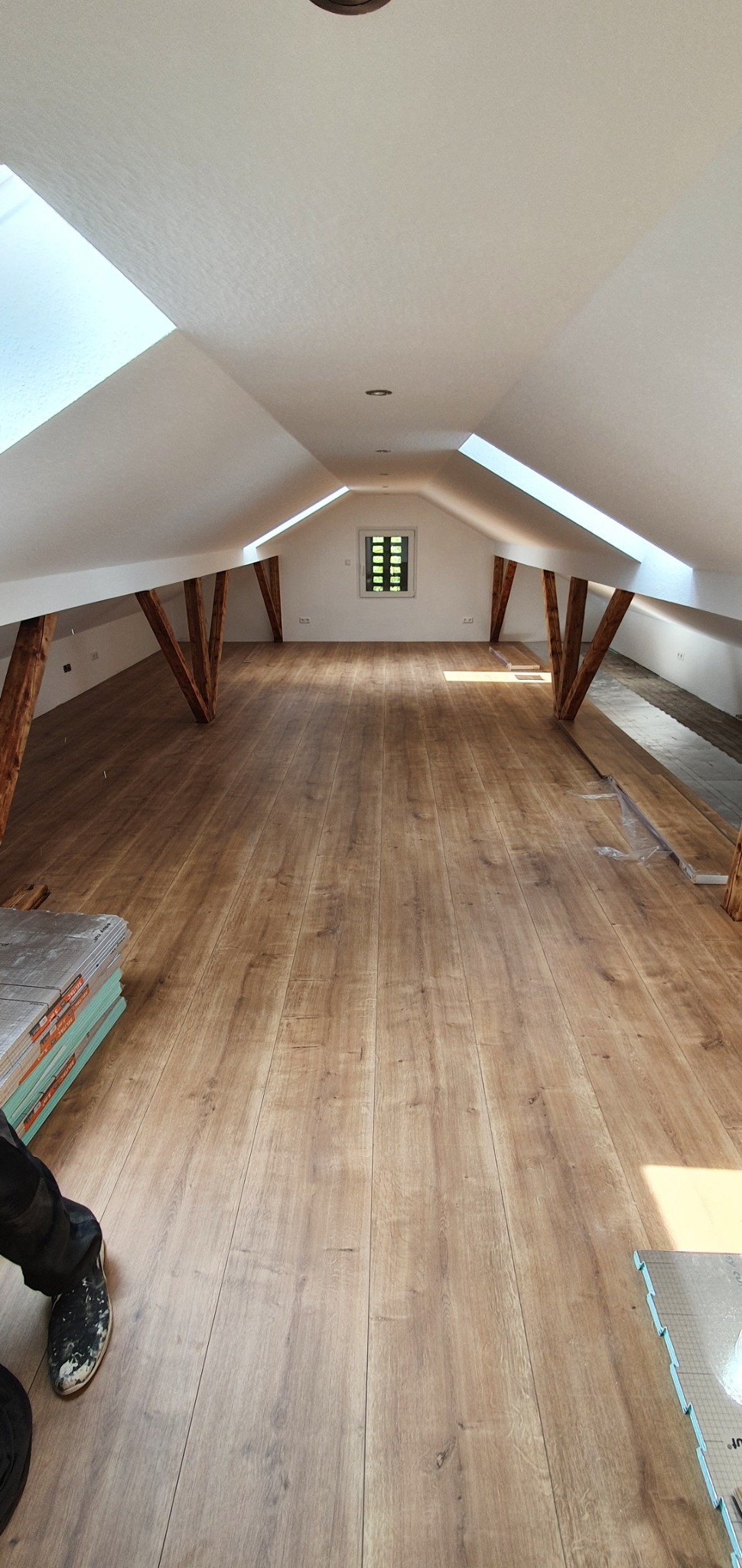 Montaż paneli podłogowych w jasnym, przestronnym poddaszu z oknami dachowymi i drewnianymi belkami nośnymi; widoczne paczki paneli i robocze buty.