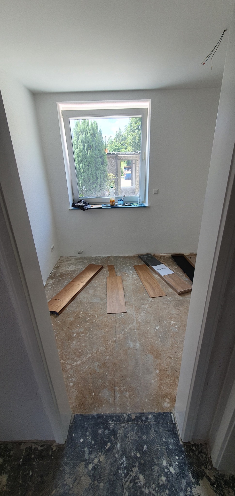 Przygotowanie do montażu paneli podłogowych w jasnym pokoju z oknem, widoczne rozłożone panele, paczka paneli i przygotowane podłoże.