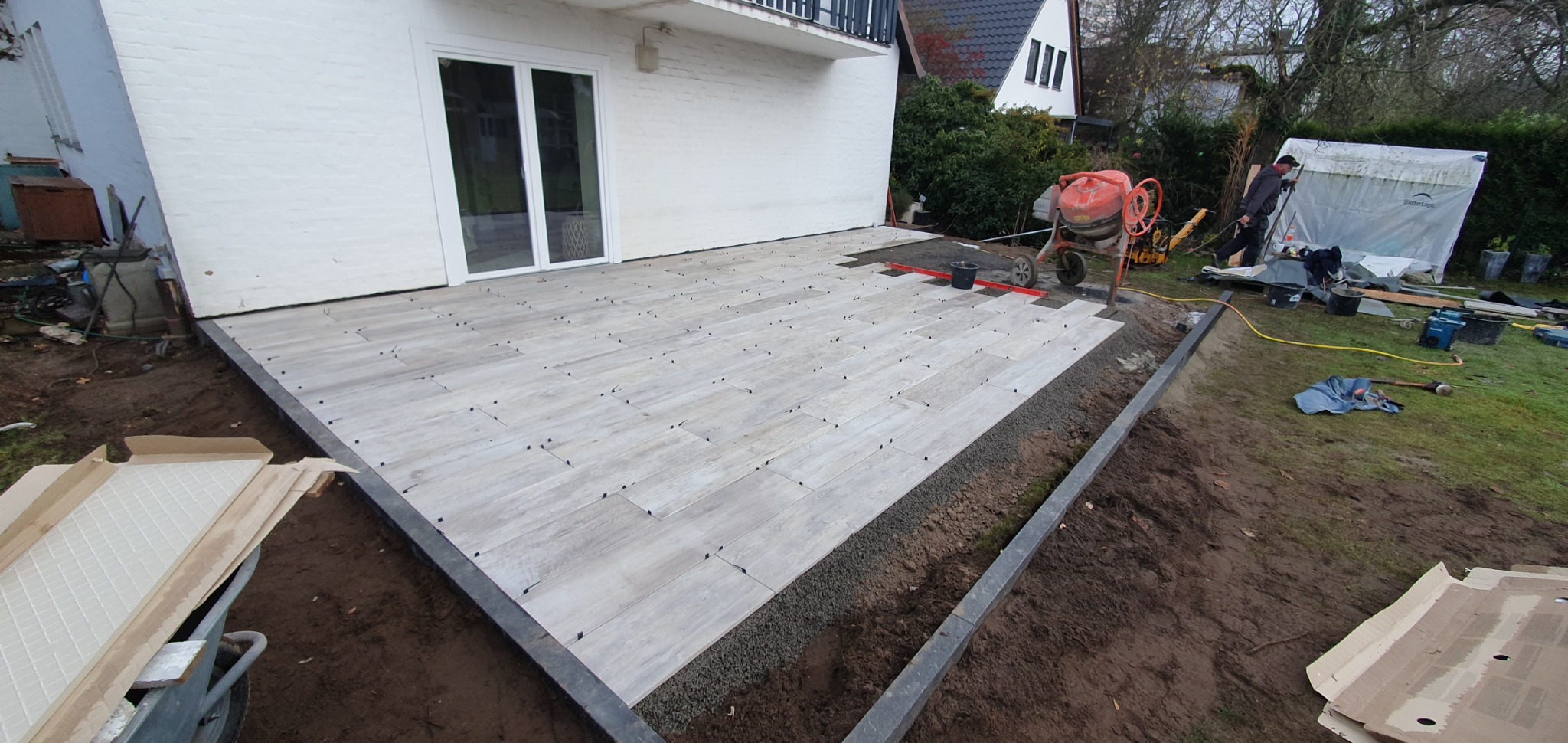Układanie płytek imitujących drewno na tarasie, widoczne czarne krzyżyki dystansowe, obrzeże z kostki granitowej, mieszarka do betonu w tle.