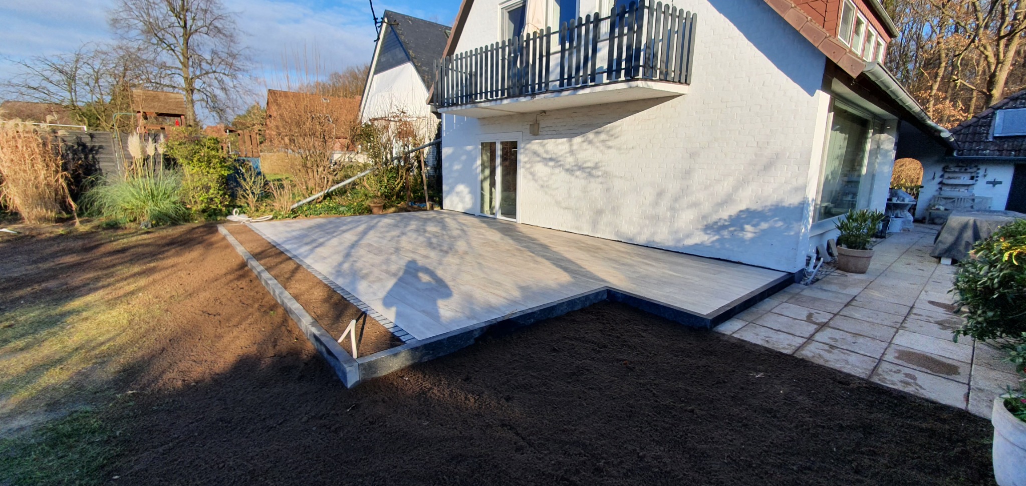 Nowoczesny taras z jasnych desek kompozytowych, obrzeża z ciemnej kostki, świeżo nawieziona ziemia pod trawnik, cień osoby fotografującej na tarasie.