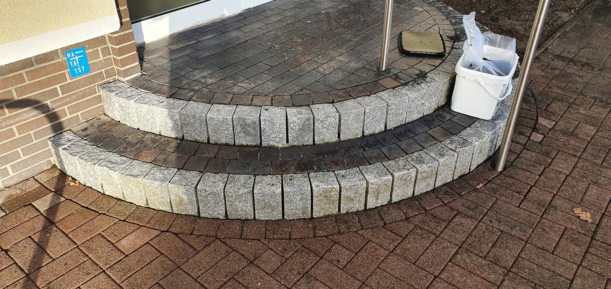 Dwu-stopniowe schody wejściowe wykończone kostką granitową i ceglaną, prowadzące do drzwi, z widoczną kostką ułożoną w jodełkę na podeście.