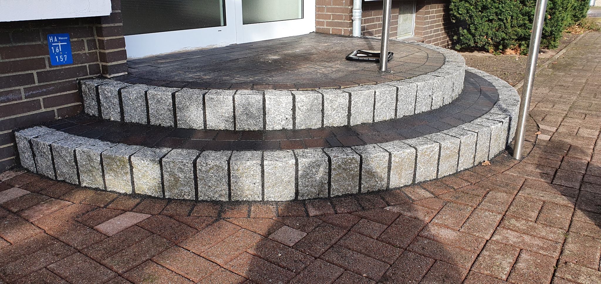 Półokrągłe, dwustopniowe schody wejściowe z kostki granitowej, prowadzące do drzwi, z kostką brukową w kolorze ceglastym wokół.