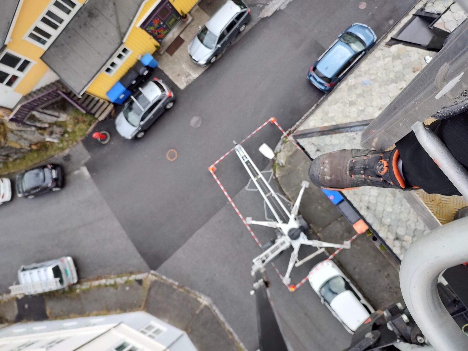 Widok z góry na zwyżkę (podnośnik koszowy) z perspektywy pracownika, w tle zaparkowane samochody i budynki mieszkalne, widoczny but roboczy na krawędzi kosza.