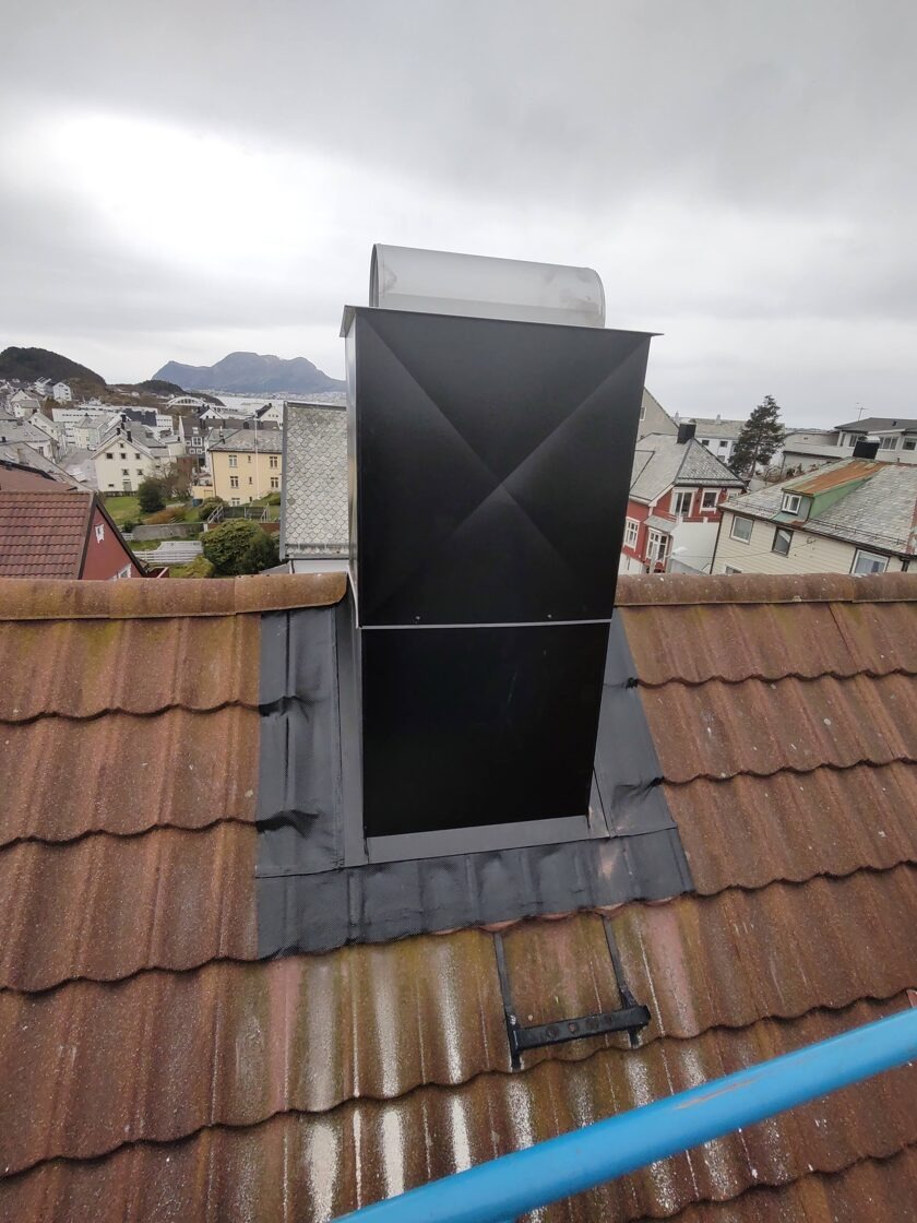 Obróbka blacharska komina na dachu z czerwonej dachówki, widoczna powlekana blacha ocynkowana na tle zabudowy miejskiej i pochmurnego nieba.